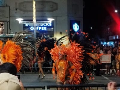 Nighttime scene captures a parade with performers in elaborate orange and black feathered costumes. They are walking on a street lined with people, some of whom are watching or taking photos. Behind them, a Starbucks Coffee sign is visible. The setting suggests a public celebration, possibly a carnival or festival. The presence of onlookers and the Starbucks indicate an urban environment.