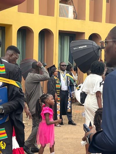 The scene depicts an outdoor gathering on a reddish-brown paved surface in Madina, Ghana, against a backdrop of a light yellow building with multiple arched window frames and an air conditioning unit on its upper level. The event appears to be a graduation ceremony or celebration, characterized by individuals wearing academic gowns and traditional Ghanaian Kente cloth sashes.
In the mid-ground, a young girl, approximately 5-7 years old, with braided pigtails, is prominently positioned, wearing a vibrant pink ruffled dress and looking to her left. Directly behind her, a man in a grey long-sleeved shirt adjusts his head or hair. To the girl's right, a woman in a black academic gown and cap, adorned with a multi-colored Kente cloth sash and a round medallion, stands smiling. Beside her, a man in a dark suit, white shirt, dark sunglasses, and a large gold medallion on a blue and yellow ribbon, also wears a Kente cloth sash over his shoulders, looking slightly to his left.
Two large black photography softboxes are visible in the mid-ground, with one labeled "Godox P120L," indicating a professional photo shoot is occurring. To the far left, a person in a black academic gown with a Kente cloth sash holds a blue folder or certificate. On the far right foreground, a person in a dark suit with spectacles holds a smartphone and a white tissue, partially obscuring the view. Further back on the right, a woman in a white dress with ruffled shoulders is seen from behind. Additional individuals, some in academic attire, are partially visible in the background.