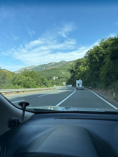 The image depicts a view from inside a vehicle driving on a winding road near Golubovići, Montenegro. The road has solid white lines delineating the lanes. In the distance, a white truck is visible on the road ahead. A mountain range covered in green vegetation is visible in the background, along with a clear blue sky with scattered clouds. Part of the vehicle's interior, including the dashboard and a small electronic device mounted on it, are in the foreground.