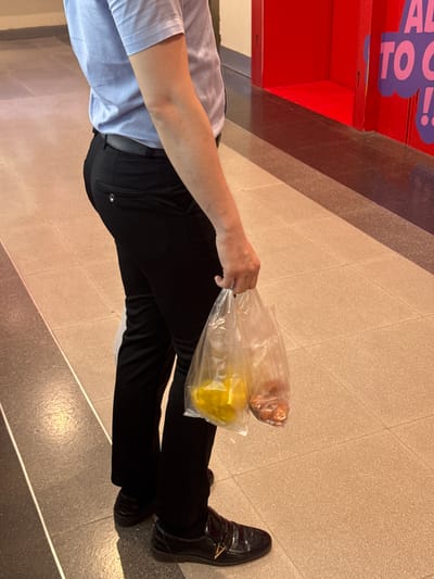 A person is standing on a light-colored tiled floor in what appears to be a public indoor area. The person is wearing a light blue short-sleeved collared shirt, black trousers, and black dress shoes. They are holding two clear plastic bags in their right hand. One bag contains yellow items, and the other contains small reddish-brown items. In the background, a bright red structure with purple lettering is visible. The scene suggests a person who has made a purchase. There is no visible information to pinpoint the location as Nong Pa Khrang, Thailand.