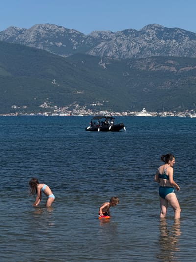 In this aerial shot, people enjoy a sunny day at the water's edge. The scene is set in Mrčevac, Montenegro, with a backdrop of a lush, tree-covered mountain range meeting a clear blue sky. A town with white buildings is nestled at the foot of the mountain, and a large white yacht is docked nearby. The water is a deep blue, reflecting the bright sunlight, creating a shimmering effect.
In the foreground, several individuals are engaged in water activities. A woman in a blue bikini stands in the water, looking to the right. Closer to the shore, a young boy, appearing to be a child, floats on an orange inflatable. Further down, another young person, also in a light blue bikini, is partially submerged in the water, performing a dive with their arms outstretched. The overall atmosphere suggests a leisurely summer day, with a sense of fun and relaxation. The reflection of the people in the water adds a unique visual element to the scene. A black boat with a canopy is visible further out on the water. The time of day appears to be midday, given the bright sunlight and the long shadows cast by the people in the water.