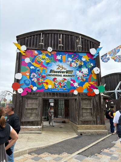 This vibrant photograph captures the exterior of a building at Universal Studios Japan in Osaka, celebrating its 25th anniversary. The central feature is a large, colorful mural covering a significant portion of the wooden facade. The mural prominently displays "25th Discover U!!!! UNIVERSAL MARKET" and "UNIVERSAL STUDIOS JAPAN", surrounded by an array of popular cartoon characters, including Hello Kitty, Minions, Snoopy, Woodstock, Elmo, Cookie Monster, Ernie, Bert, Big Bird, and Woody Woodpecker. These characters are depicted in dynamic, celebratory poses amidst colorful abstract shapes, clouds, and lightning bolts, creating a festive and energetic visual.

The building itself features dark wooden siding with a curved roofline and two multi-paned windows to the right of the mural. Three decorative light fixtures are mounted on the wooden wall. A sign above one window reads "ROY BARNES," likely the name of the establishment.

In the foreground, a paved walkway or street is visible. Several people are present: a man and woman are partially visible in the bottom right, looking downwards. Further back, near the building's entrance, another woman in light-colored clothing stands, looking left. Against a bright, overcast sky in the upper left, several butterfly-shaped banners bearing "25th" are suspended, reinforcing the anniversary theme. The scene conveys a lively daytime atmosphere at a theme park, marking a special event or milestone.