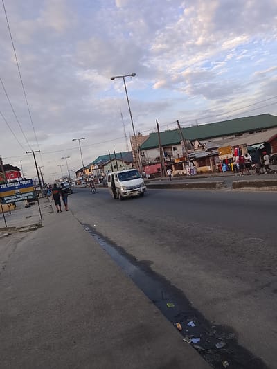 The image shows a street scene in Rumuepirikom, Nigeria. The sky is overcast with scattered clouds. In the foreground, the road surface is asphalt, with a drainage ditch running along the right side, containing some debris. On the left side of the road, there is a sidewalk with a sign that reads "THE NIGERIA POLICE DIVISIONAL HEADQUARTERS RUMUEPIRIKOM PH.". Two individuals are walking on the sidewalk, facing away from the camera. A white van is driving on the road. Beyond the road, there are buildings, some with green roofs, and shops displaying clothing. Several streetlights are visible along the road. The overall impression is of a busy urban street.