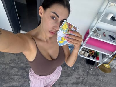 A female subject is captured in a selfie-style photograph, standing in what appears to be an outdoor or semi-outdoor area within the city of Cam Đức, Vietnam. She has dark hair, wears a brown tank top, and light pink loose pants. Visible freckles are present on her face. Her left upper arm bears a tattoo, and her right forearm displays the text "TU CORVO TUTTO." Her fingernails are painted pink with decorative elements. The subject holds four tarot cards fanned in her right hand, facing the viewer. The clearly visible cards, from top to bottom, are "The Sun," "The Moon," and "The Star." A fourth card, partially obscured by her thumb, is at the bottom. The immediate environment includes a grey concrete floor. To the subject's left, a dark doorway or opening is visible. To her right, a white metal shelving unit with multiple tiers is present. The shelves contain miscellaneous items: the top shelf holds a white roll and a dark object; the second shelf contains several metallic discs, a decorative shell, a red multi-tool or lighter, and a small black object; the third shelf features a rolled pink mat and a clear plastic bottle; and the fourth shelf displays multiple pairs of shoes. A yellow broom is situated on the concrete floor beneath the shelving unit. The background wall is white.