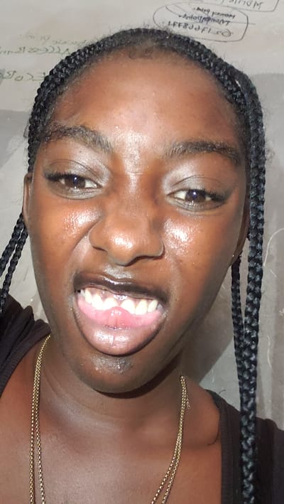 The media file is a close-up, low-angle, headshot of a young Black female with braided hair. She is looking directly at the camera with a wide, toothy smile, her mouth open, revealing her teeth and tongue. Her eyes are open, and her gaze is directed forward. She is wearing a dark top and a delicate gold chain necklace.
The background appears to be a wall with some faint, handwritten text in what looks like English script. One legible portion of text in a circular outline reads, "NDUISE", followed by "Nudged". There are other faint scribbles that are difficult to decipher. The lighting suggests it might be indoors or an overcast day, as there are no strong shadows. The setting does not provide many specific cues about the location beyond the text on the wall, which, when combined with the provided location context of Tudun Nupawa, Nigeria, suggests the image was taken there. The subject's expression conveys happiness and a sense of joy. The overall impression is a candid, self-taken photograph capturing a moment of cheerfulness.