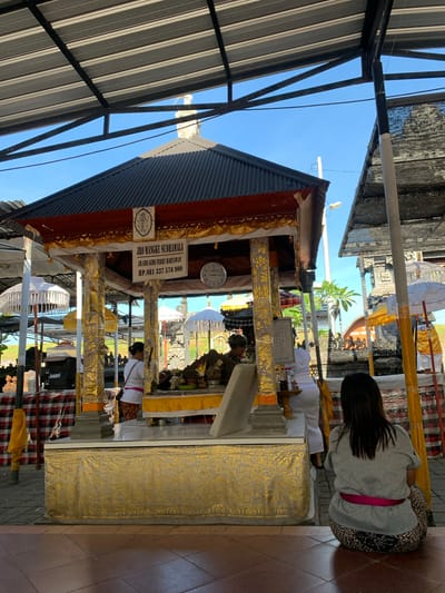 In Buleleng, Indonesia, this image depicts a scene at a Hindu temple. The foreground shows the back of a woman seated on tiled steps, facing away from the viewer. She is wearing a grey t-shirt and patterned pants with a pink sash. Behind her, an elevated structure, likely an altar or shrine, is adorned with gold fabric and pillars. Two women in traditional Balinese attire, one in white and the other in a white top and patterned sarong, are attending to offerings at the shrine. A sign above the shrine displays text, including what appears to be a name and phone number. A round clock is visible on the right side of the shrine. Several white and yellow umbrellas, typical of Balinese temple decorations, are placed around the area. The background shows more temple architecture and lush greenery under a clear blue sky, suggesting an outdoor setting. The overall ambiance is one of religious observance and cultural heritage.