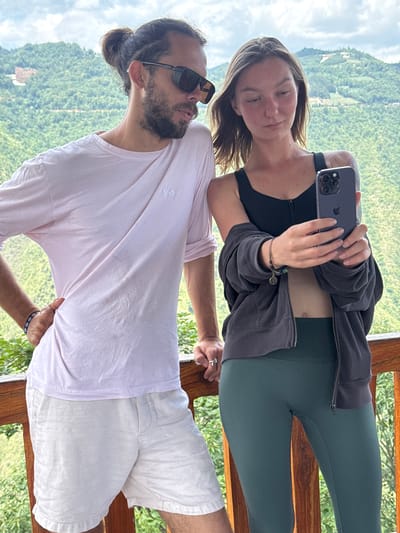 Two people stand on a wooden balcony overlooking a lush green landscape. A woman holds a smartphone, possibly taking a selfie, and leans against a man. The woman wears a black sports bra, a dark jacket, and dark green leggings. The man wears sunglasses, a pink shirt, and white shorts. The background reveals a green, mountainous terrain with hints of buildings, suggesting a location in or near a populated area. The scene is set during the daytime with natural light. The location is likely Lũng Cú Commune, Vietnam, based on the geographical description and identifying cultural characteristics.