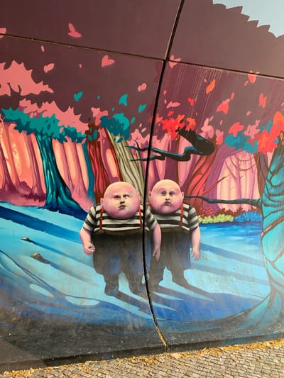 This is an outdoor mural painted on a curved surface, possibly a bench or a decorative element, set against a cobblestone ground. The mural depicts a surreal, dreamlike scene with two pale-skinned, bald figures reminiscent of infants, wearing striped shirts and suspenders. Their faces have a slightly bewildered or unnerving expression. One figure lies on its back with arms outstretched, while the other is positioned similarly, with only its head and torso visible.
The background is a vibrant and abstract landscape. To the left, there's a dark, stylized tree with red and pink leaves, set against a purple backdrop. To the right, a blue, watery landscape with abstract shapes and reflections is depicted. The overall style is painterly and stylized, with bold colors and fluid lines.
The image is taken from a slightly elevated angle, and the mural is divided into sections by visible seams or cracks. The lighting suggests it is daytime, with no indication of specific weather conditions beyond what the natural sunlight illuminates. There are no visible text elements in the mural or its surroundings. The setting appears to be a public space, given the cobblestone pathway. The surreal and unsettling depiction of the figures, combined with the fantastical landscape, creates a mysterious and perhaps slightly disquieting atmosphere.