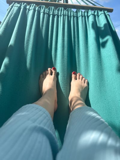 A first-person perspective shot captures two bare feet, each with red nail polish, resting on a teal-colored fabric hammock. The hammock is suspended by a wooden bar and ropes, and sunlight casts dappled shadows across its surface. The person appears to be wearing light blue, ribbed pajama pants. The background consists of a clear blue sky and hints of greenery, suggesting an outdoor setting. The image was taken in Vyshevychi, Ukraine.