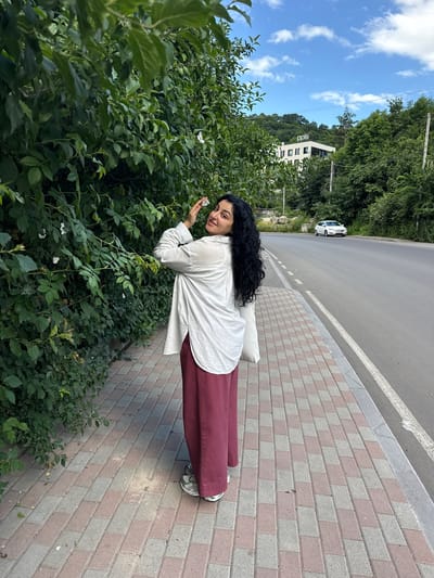 A young woman with long, dark, curly hair stands on a paved sidewalk next to a dense green hedge. She is wearing a loose-fitting white shirt and long, wide-leg burgundy pants. Her right arm is raised, and she is touching a white flower in the hedge with her fingers. She is looking over her left shoulder at the camera with a slight smile. Behind her, a road stretches into the distance, with a white car driving on it. Further in the background, a modern white building with the letters "ppls" is visible on a hillside, surrounded by lush green trees. The sky above is blue with scattered white clouds. The sidewalk is made of interlocking rectangular bricks in shades of gray and pink.