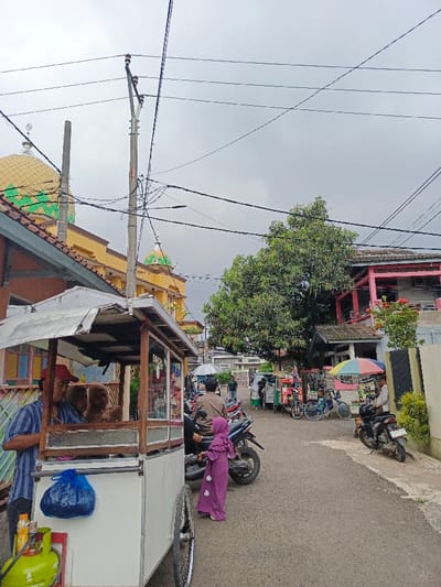 A bustling street scene features a food cart with two vendors, a yellow mosque with green domes, and power lines crisscrossing the cloudy sky. A small child in a purple dress stands near a parked motorcycle, while other pedestrians and vendors populate the street. Various bicycles and motorcycles line the road, and several small businesses operate on the sides of the street. A large tree adds shade to the scene.