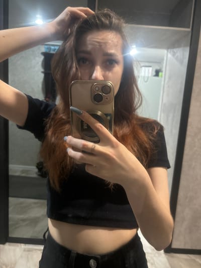 This media file shows a young woman taking a mirror selfie in what appears to be a bathroom or dressing room. She has long, reddish-brown hair that she is touching with her left hand, and she is looking into the camera with a slight frown or pensive expression. She is wearing a black crop top and dark-colored pants, possibly jeans. Her nails are painted a light blue color.

She is holding a smartphone in her right hand, which is positioned to capture her reflection. The phone case is light beige with a gold camera bump. A thin, gold ring with a twisted design is visible on her right ring finger.

The reflection in the mirror shows the back of the room, which has light-colored, possibly tiled walls. The lighting appears to be artificial, suggesting it could be daytime or nighttime. The overall setting feels personal and private, consistent with a selfie taken in a home or changing area. There is no visible text in the image. The orientation of the image is landscape, with the subjects appearing sideways.