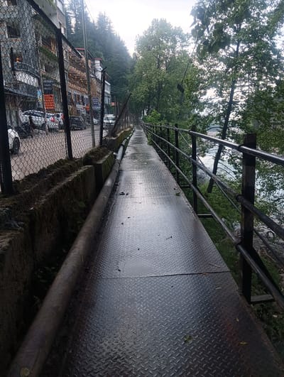 The image captures a narrow metal walkway beside a flowing river, likely in Manali, India. The walkway, made of diamond plate metal, appears damp, possibly from recent rain, reflecting the overcast sky. A black metal railing runs along the outer edge of the walkway, separating it from the river. To the left of the walkway, a chain-link fence encloses a street lined with parked cars and buildings. The buildings, with their balconies and signage, suggest a commercial area. Dense trees and foliage are visible on the opposite bank of the river and on the hillside behind the buildings. The overall atmosphere is serene and natural, with a hint of urban development along the riverfront.