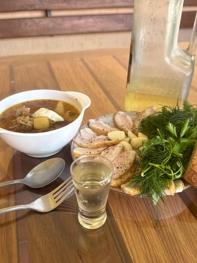 A close-up shot captures a table laden with a traditional Ukrainian meal. In the foreground, a white bowl brimming with hearty soup, likely borscht, sits on a wooden table. Alongside the soup, a plate displays thinly sliced salted pork belly, seasoned with pepper, and interspersed with cloves of garlic. A bunch of fresh dill and parsley garnishes the plate, along with a slice of rustic bread. A shot glass filled with a clear liquid, presumed to be vodka, stands in front of the plate. To the left, a spoon and fork rest on the table. In the background, a large glass decanter, partially filled with a yellow liquid, hints at a beverage. The scene is set on a wooden table with a backdrop of wooden slats, suggesting an outdoor or rustic indoor setting. The overall composition evokes a sense of comfort and hospitality, typical of a home-cooked meal. The image is devoid of human presence, focusing solely on the culinary spread. The presence of a wooden table and rustic elements could suggest a location like Hraniv, Ukraine, where such traditional meals are commonly enjoyed in a relaxed, domestic setting.