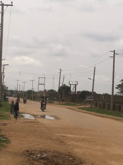 The image shows a dirt road in Hanwa, Nigeria. Several people and motorcycles are on the road. Overhead power lines run along the roadside. Some puddles of water are visible on the road. The sky is overcast. Buildings and vegetation are visible on the side of the road. A metal structure resembling a water tower can be seen further down the road.