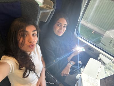 Two women are posing for a selfie on a train. One is wearing a white shirt and the other is wearing a black outfit and headscarf. A laptop, water bottle, and purse are visible on the table in front of them. The background shows a passing landscape through the train window. It's not possible to determine the specific location in Sion, Switzerland from the image.