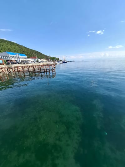 This media file captures a scenic view of a body of water, likely Lake Baikal given the location context of Listvyanka, Russia. The water is a deep, clear turquoise, allowing visibility of the seabed in shallower areas. A wooden pier extends into the water, supporting a series of industrial-looking structures, possibly related to fishing or aquaculture. Along the shoreline, there's a verdant, tree-covered hillside dotted with buildings, including a white building with a bright blue roof and a Ferris wheel in the distance, suggesting a tourist or recreational area. The sky is a clear, bright blue with a few wispy white clouds. The sun appears to be high, indicating it is daytime, likely mid-morning or early afternoon, under fair weather conditions. There are no people explicitly visible in the frame, though the presence of recreational facilities hints at human activity. The overall impression is one of a serene, picturesque location with a blend of natural beauty and human development.