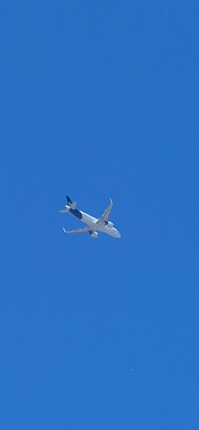The image captures an airplane flying in a clear blue sky. The airplane is white with a dark tailfin that features a logo. It appears to be a commercial passenger jet. The perspective suggests the photo was taken from below, looking upwards. There is no discernible urban landscape or any specific landmarks that would indicate the city of Mainz, Germany. The environment is solely the sky.