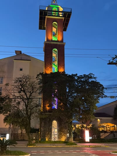 The image shows a tall, illuminated clock tower at dusk in Florianópolis, Brazil. The tower, made of brick and stone, is lit with orange and green lights, highlighting its architectural features. A spiral staircase is visible inside, also lit with green light. The tower is surrounded by trees and other buildings, including a light-colored structure to the left and a restaurant with outdoor seating to the right. Power lines crisscross the sky. The scene suggests a public space, possibly a plaza or a street corner, where the tower serves as a prominent landmark.