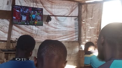 Four men are sitting in a room watching a soccer game on a flat screen television mounted on the wall. The room is sparsely furnished with wooden walls and a corrugated metal roof. The television is displaying a soccer match with players in red and white jerseys. The men are all wearing casual clothing. One of the men is wearing a blue shirt with the word "OTE" on the back. Another man is wearing a blue shirt with the word "OTE" on the back. Another man is wearing a light blue and yellow striped shirt. The fourth man is wearing a light blue and green shirt. The television is hooked up to a DVD player on a small table in the room. There is a window with a wooden frame and a broken pane in the back wall of the room. Sunlight is streaming through the window. The scene is located in Ota, Nigeria, at the coordinates 6.69 latitude, 3.177 longitude.