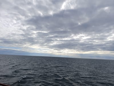 The image depicts a wide expanse of the ocean under a cloudy sky. The sea is a dark, choppy blue, with small waves creating whitecaps on the surface. The horizon is visible as a sharp line separating the water from the sky, which is filled with a variety of gray and white clouds. Some of the clouds are thin and streaky, while others are thicker and more textured. Sunlight appears to be breaking through in some areas, creating brighter patches in the sky. In the foreground, a dark, reddish-brown railing or edge of a structure is partially visible along the bottom left of the frame, suggesting the image was taken from a boat or a pier. The location is described as Unknown, Sweden.