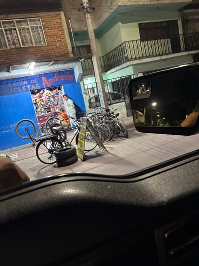 The image depicts a nighttime street scene in León, Mexico, viewed from inside a vehicle. The foreground is dominated by the dark dashboard and a partial view of a hand or arm on the lower left. A side mirror on the right reflects a dimly lit street with multiple streetlights and dark skies.
The primary focus is a bicycle repair shop named "Andrade" with a bright blue facade. The shop's sign, "REPARACIÓN DE BICICLETAS Andrade," is visible in white and red lettering. The interior of the shop is brightly illuminated, revealing a dense display of bicycle parts and accessories. Three individuals are present near the shop's entrance: a man in a light-colored shirt stands outside, facing generally towards the camera. Inside the shop, two other individuals are partially visible, one in a darker shirt and another wearing an orange garment, both appearing to be seated or leaning.
On the sidewalk in front of the shop, a collection of bicycles is parked. One bicycle is notably positioned upside down, resting on a stack of two black tires. A yellow A-frame sign stands upright near this bicycle. Additional bicycles are lined up along the building facade further to the right.
The surrounding environment consists of urban buildings. Above the bicycle shop, a brick building with gridded windows is visible. To the right of the shop, a two-story building features a light green/teal facade with dark wrought-iron balconies and window grilles. A utility pole with multiple wires stands centrally on the sidewalk. The overall lighting is artificial, emanating from the shop's interior, streetlights, and ambient city glow.