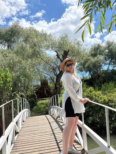 A young woman stands on a curving wooden pathway or bridge, looking towards the camera with a subtle smile. She has blonde hair and is wearing a straw hat, dark sunglasses, a loose white long-sleeved shirt over black shorts, and white sneakers. Her right hand rests lightly on a white metal railing that follows the curve of the path.
The scene is an outdoor setting, likely a park or natural area, characterized by abundant greenery and a bright sky. Above and around the pathway, numerous trees with light green, leafy foliage are visible. Some long, narrow green leaves hang into the top-left of the frame, hinting at an overhead tree, possibly a willow. The sky is a clear blue with scattered white, puffy clouds, indicating sunny weather during the daytime. Below the pathway and railing, lush green vegetation, possibly reeds or tall grass, grows alongside what appears to be calm, reflective water. The wooden pathway itself features parallel planks, suggesting a well-maintained scenic route. No text is visible in the image. The atmosphere is relaxed and pleasant.