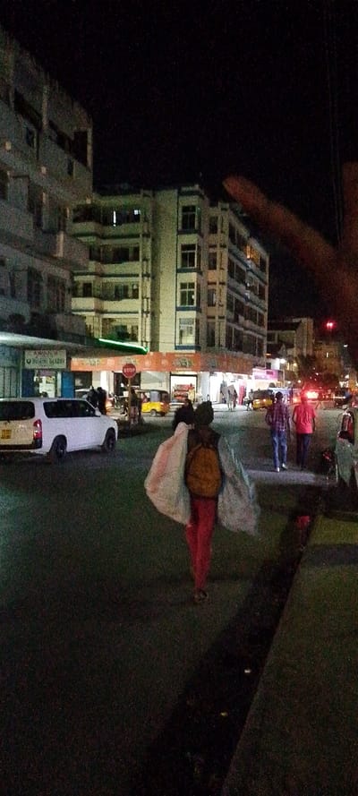 The image depicts a nighttime street scene within an urban area of Mombasa, Kenya. The foreground features the back of an individual walking on a paved road surface, wearing a dark top, red pants, and an orange backpack, carrying two large, light-colored bundles, one over each shoulder. To the right of this individual, two other male pedestrians walk away from the camera; one wears a plaid shirt and blue jeans, the other a red shirt and dark pants.

On the left side of the road, a white Toyota Probox van is parked. In the mid-ground, a red octagonal "STOP" sign is visible. Multi-story buildings with illuminated shopfronts line both sides of the street. One visible establishment on the left is labeled "PROMOTE BAKERY." Further down the street, at least two yellow tuk-tuks are present. The environment is illuminated by artificial streetlights and commercial lighting from the shops, set against a dark night sky. A blurry, reddish shape, consistent with a finger or hand, partially obscures the upper right corner of the frame.