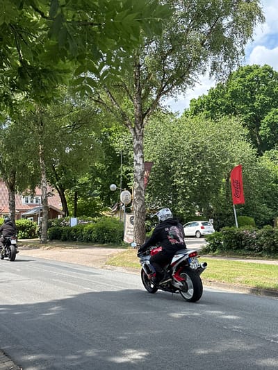 Two individuals on motorcycles are observed from behind, traveling away from the viewer on a paved road in Oberndorf, Germany. The scene depicts a tree-lined street under a partly cloudy sky.
The lead motorcyclist, positioned further down the road on the left, rides a dark-colored motorcycle. This rider wears a dark helmet and a dark jacket or hoodie. The motorcycle displays a German license plate reading "STD M 10".
The second motorcyclist, closer to the foreground and slightly to the right, operates a red and white sportbike with a white rear wheel and a silver exhaust pipe. This rider is wearing a white motocross-style helmet with a visible "FOX" brand logo on the back, and a black hoodie featuring a multi-colored graphic print of what appears to be an anime-style female character on the back. The motorcycle bears a German license plate stating "STD OS FI 44".
The road is asphalt, appearing dry with some visible tire marks and shadows cast by unseen objects to the left. A dirt and grass verge separates the road from the surrounding environment on the right.
To the right of the road, various signs and structures are visible. A red flag with the white "Coca-Cola" logo and the German text "WERBUNG / ERFRISCHT" is mounted on a pole. Further along, a small round sign resembling a traditional German inn or brewery emblem is affixed to a pole, below which is a black rectangular sign with white text advertising "Parkplatz für Gäste" (Parking for Guests) and "Crêperie". A white "P" (Parking) sign is also visible behind the second rider.
The background consists of lush green deciduous trees and bushes. Several houses with red tiled roofs and white facades are partially visible through the trees on the left side of the road. A white compact car is parked further back on the right, partially obscured by foliage. The overall environment suggests a suburban or semi-rural setting with established greenery.