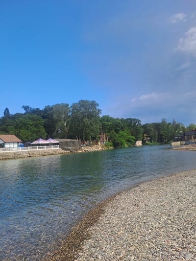 The image depicts a scenic view of a riverbank in Dzhubga, Russia. The foreground is dominated by a pebble beach that slopes gently into the clear, blue-green water of the river. The water is calm, with subtle ripples reflecting the clear blue sky above. Along the river's edge, on the left side of the frame, is a white, low-lying building with a brown roof, partially obscured by trees. Adjacent to this building is a white railing, and behind it, two purple umbrellas are visible, suggesting a seating area or cafe. Further along the riverbank, a dense line of lush green trees forms a backdrop, with some taller trees interspersed among them. The river curves to the right, leading the eye towards more distant structures and vegetation. The overall scene is tranquil and bathed in natural light, with a few wispy clouds present in the upper right portion of the sky. There are no people visible in the image.