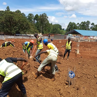 This media file depicts an active construction site in Borabu Chitago ward, Kenya, under a bright, partly cloudy sky. At least six male laborers are engaged in manual groundwork, primarily digging and moving large quantities of reddish-brown soil.
The scene is dominated by the excavated earth, indicating the early stages of a building foundation. Workers are seen using various shovels with orange, blue, and green handles to break up and displace the soil. Most individuals wear practical work clothes, including bright yellow or lime green high-visibility vests, and some are equipped with protective hard hats (one orange, one blue). One worker's vest in the foreground displays partial text, "GROUP."
Scattered across the site are several concrete foundation pads with exposed rebar, signifying the structural elements being prepared. A blue hard hat is visible resting on a rebar cage in the lower right of the frame. In the background, numerous green trees line the horizon, and a structure with a distinct light blue corrugated metal roof is visible, suggesting a nearby existing building or a future part of the development.
The clear sky and strong shadows indicate it is daytime, likely mid-morning or early afternoon. The workers appear focused on their demanding physical tasks, moving earth to prepare the site for further construction. The overall impression is one of diligent manual labor in a sunny, semi-rural construction environment.