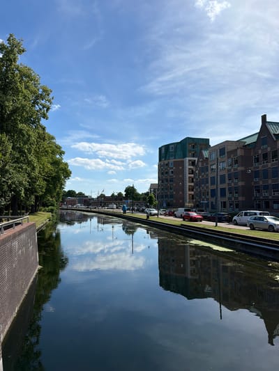 The image depicts a canal in Helmond, Netherlands, on a sunny day with scattered clouds. The perspective is from above and to the side of the canal, with the scene oriented vertically, appearing upside down.
On the left side of the image, a lush green treeline runs along the edge of the canal. Further beyond the trees, modern buildings with numerous windows and dark facades line the opposite bank. Cars are parked along the street in front of these buildings. The reflection of the buildings and the sky is clearly visible on the still water of the canal. A brick walkway or path is visible in the foreground.
The sky is bright blue with wispy white clouds, suggesting it is daytime, likely late morning or early afternoon. The clear sky and visible reflections indicate fair weather. There is no discernible activity or event taking place other than the general ambiance of a town or city alongside a waterway.
Visible text includes "aspa" on a white car parked on the side of the road. Other text is too small or indistinct to read. The overall mood of the image is serene and captures a typical day in this European location.