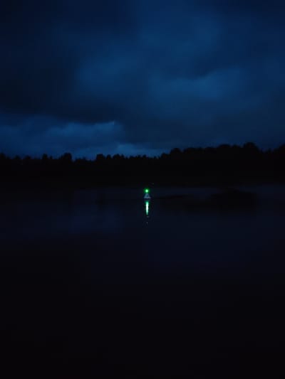 This is a nighttime photograph of a lake in Devyatiny, Russia. The sky is dark and cloudy, with a hint of blue. A dark line of trees forms the horizon across the middle of the image. In the foreground, the water is dark and reflective. In the center of the lake, a navigational buoy with a green light is visible. The buoy is white and conical with a green light at its peak, and its reflection is visible on the water. The scene is dimly lit, creating a serene and slightly mysterious atmosphere.