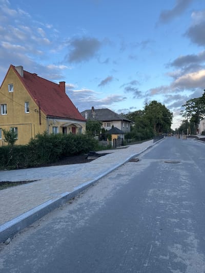 The image depicts a street scene in Guryevsk, Russia, during what appears to be late afternoon or early evening. The sky is partially cloudy with a mix of blue and grey.
In the foreground, a new asphalt road is visible, with a newly constructed sidewalk or pedestrian path on the left. The sidewalk is made of light-colored paving stones and bordered by a granite curb. Adjacent to the sidewalk, there is a strip of soil and some landscaping materials.
Several houses are visible along the street. On the left, a two-story yellow house with a red tiled roof and white-framed windows stands out. Further down the street, other houses with pitched roofs can be seen. Some trees and dense green bushes line the property borders.
The road itself appears to be undergoing some form of construction or renovation. There are manhole covers visible in the asphalt. In the distance, there are some construction materials, including pallets and possibly yellow construction equipment. Streetlights and utility poles are present along the side of the road. The overall impression is of a residential area in transition, with infrastructure improvements underway. There are no people visible in the image.