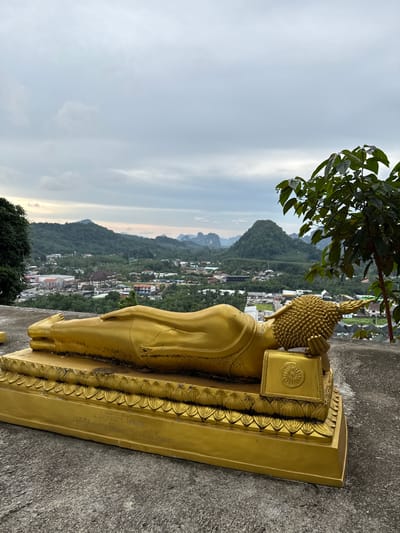 In Ban Ao Nang, Thailand, a golden, reclining Buddha statue lies on a platform. The statue is positioned in the foreground, facing right, with its head resting on its arm. Behind the statue, a cityscape unfolds with buildings and lush green hills dotted with karst formations. The sky above is overcast with clouds. A tree with dark green leaves is visible on the right side of the image, partially obscuring the view of the city and hills. The base of the statue has inscriptions in an unfamiliar script.
