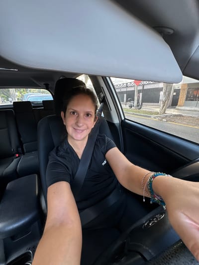A woman wearing a black shirt and a seatbelt is sitting in the driver's seat of a car. She is holding the steering wheel and smiling at the camera. The car appears to be in Miraflores, Peru, as evidenced by the architectural style of the buildings visible through the windshield. A parked scooter is visible outside. The interior of the car is black. The woman has multiple bracelets on her right wrist.