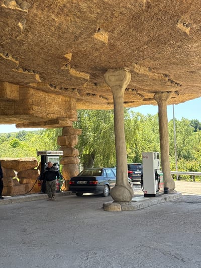 The image shows a gas station in Belomortsi, Bulgaria. The gas station features architectural elements resembling large, stylized bones attached to a textured, earth-toned wall. A black car is parked at one of the pumps, and a man wearing a dark jacket and camouflage pants is standing next to the pump, appearing to be filling up the car. The surrounding environment includes trees and other vehicles, suggesting a typical roadside gas station setting. The weather appears sunny and clear.