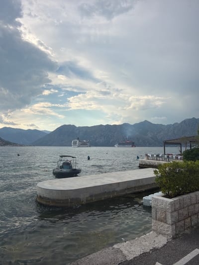 In Dobrota, Montenegro, a serene seascape unfolds under a dramatic sky of billowing clouds. The water, a deep, reflective gray, laps gently against a concrete pier in the foreground, where a small, dark inflatable boat with a canopy is docked. To the right, a stone retaining wall borders a patch of lush green foliage, with tables and chairs set up under a pergola, suggesting an outdoor dining area.

In the middle ground, two large cruise ships are visible on the expansive bay. The ship on the left is white with red accents, while the one on the right is predominantly white with red and black detailing and a plume of smoke rising from its stack. In the distance, imposing, dark mountains form a majestic backdrop, their peaks softened by the atmospheric haze. The scene is quiet and still, with no visible people, hinting at a peaceful moment captured in time.