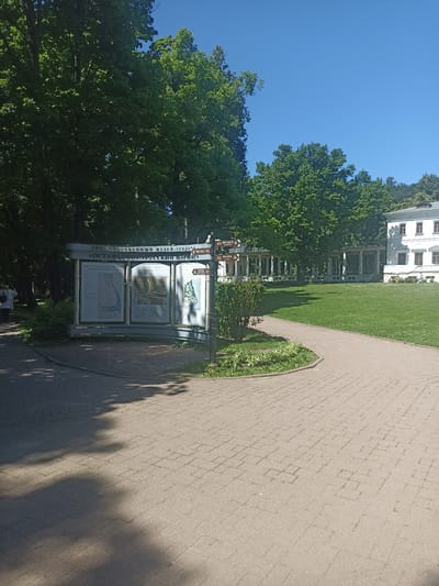 An exterior shot captures a sunny day in Ostafievo, Russia, with a clear blue sky above lush green trees. In the foreground, a paved pathway, made of tan interlocking bricks, curves to the right. To the left of the pathway stands a curved informational kiosk with multiple display boards, featuring maps and text in Russian. A lamppost with directional signs points towards "MUSEUM" and "CAFE." Behind the kiosk, a cluster of green bushes and some plants add to the natural scenery. Beyond the bushes, a grassy lawn stretches out, leading to a long, white building with a colonnade. The building has several windows with brown frames and a visible roofline. The overall scene suggests a park or historical estate, likely a tourist attraction.