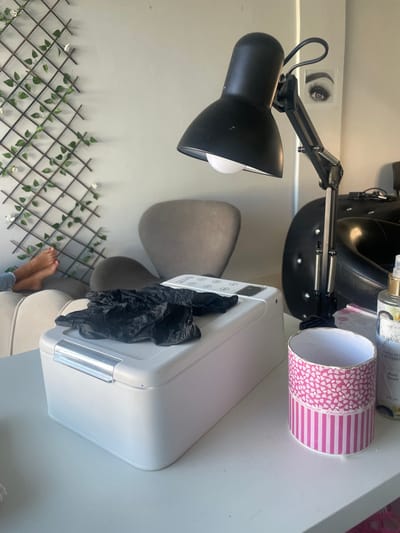 The scene is set in an interior room, likely a beauty salon or spa, in Anápolis, Brazil. The foreground features a white table. On the table, to the left, rests a white, rectangular electronic device with a silver front latch and a control panel on its top surface. A pair of crumpled black nitrile gloves is placed directly on this device. To the right on the table, a black, articulated desk lamp with a white bulb is positioned, casting light downwards. Further right on the table, a pink and white cylindrical container with a heart pattern at the top and vertical stripes below is visible. Behind this container, a clear bottle of "O BOTICÁRIO BOTANIC ORCHIDÉE NOIR Body Splash" stands.

In the midground, the bare feet of an individual, seemingly resting, are visible on a light-colored armchair or sofa to the left. A dark gray armchair with a high, rounded back is centrally located. To the far right, part of a black, possibly studded, treatment chair is visible. The background wall is light gray. On the left wall, a decorative trellis adorned with artificial green leaves and small white flowers is mounted. On the right wall, a framed image or poster depicting a stylized eye with long eyelashes is displayed. The environment appears clean and well-lit, suggesting a professional establishment.