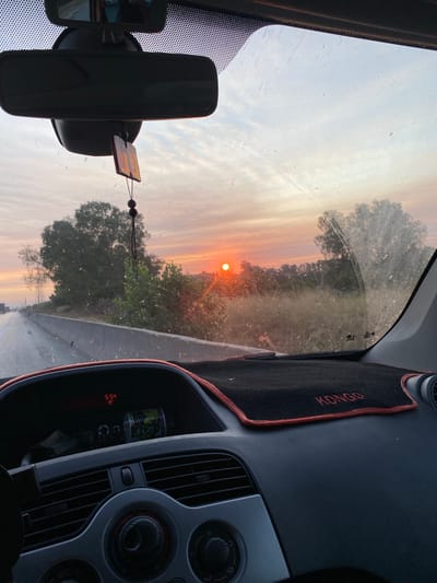 The image is a photograph taken from inside a vehicle, looking forward through the windshield. The sun is setting, casting a warm orange glow across the sky and illuminating a rural landscape. Trees and dry grass are visible on either side of a road, suggesting the vehicle is in motion. The interior of the car is also visible, including the dashboard, rearview mirror, and a hanging air freshener. A digital display on the dashboard shows the time as 5:54 and the temperature as 19 degrees Celsius. The overall scene depicts a peaceful sunset drive through the countryside. The location is identified as Beni Tamou, Algeria, which is consistent with the rural setting and the warm, dry climate suggested by the vegetation and lighting.