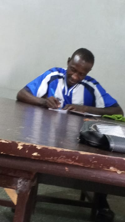An indoor scene in Dakata, Nigeria, depicts a dark-skinned male individual with short, dark hair, seated at a worn wooden table. He is wearing a short-sleeved shirt with horizontal blue and white stripes. The individual is actively writing on a small, light-colored piece of paper using a pen held in his right hand, with his head tilted downwards towards the paper. A slight smile is visible on his face. To his right, on the table, a dark-colored bag with a clear or mesh panel and a folded green fabric item are present. The wooden table displays significant wear, including chipped edges and surface scratches. The background consists of a plain, light-colored wall.
