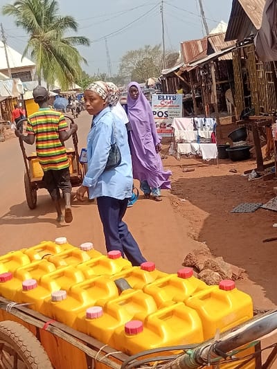 A street scene shows a cart loaded with numerous yellow jerrycans, each capped with red. Beside it, two women in headscarves, one light blue and one lavender, stand on the street. A man in a striped shirt is pulling a cart with small wheels. Businesses and merchandise are visible on the side of the street.