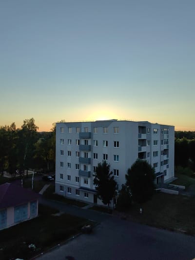 An aerial, eye-level shot captures a modern, pale gray, multi-story apartment building in Myadzyel, Belarus, bathed in the warm glow of a setting sun. The sky transitions from a pale blue at the top to a soft yellow and orange hue near the horizon, indicating either dawn or dusk. The apartment building, with its numerous white-framed windows and balconies, occupies the center of the frame. To the left, a smaller, single-story building with a dark red roof is visible. Surrounding the buildings are lush green trees, some tall and slender, others with fuller canopies. A paved road runs in the foreground and extends towards the apartment building, with a dark-colored car parked near it. The overall scene is one of tranquil urban landscape at the cusp of day or night.