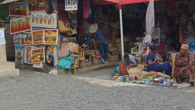 The image depicts an outdoor market scene in Moshi, Tanzania. The foreground consists of light grey gravel. In the midground, various colorful handcrafted items, including small ceramic cups and bracelets, are displayed on a dark fabric mat on the ground.
Four individuals are present within the scene. On the left, a man wearing a striped shirt and patterned fabric is seated on the ground, actively weaving a light-colored basket or mat. Next to him, a woman in a blue patterned dress sits on a blue cushion, handling small craft items. To her right, another woman in a vibrant patterned dress and headwrap is seated on a low surface, observing towards the right side of the frame. Further back and to the right, a man in a blue t-shirt and jeans stands inside the shop entrance, leaning against a display.
Behind these individuals is a shop entrance covered by a red awning, supported by a red pole with a grey folded umbrella attached. The shop's left exterior wall is adorned with multiple vibrant paintings depicting African wildlife (elephants, giraffes) and Maasai figures. A white sign displaying "ENTRY" in black capital letters is visible above these paintings. The shop interior is densely packed with various merchandise, including woven baskets, fabric goods, and other crafts. Near the left side of the shop entrance, stacked cardboard boxes, small wooden stools, and a green bag are visible.