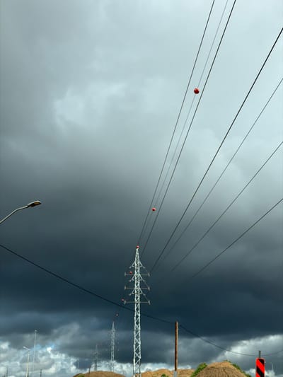The image displays an outdoor environment within the Hevel Yavne Regional Council, Israel, under a predominantly dark, heavily overcast sky. The upper portion of the sky is lighter grey, transitioning abruptly to dense, dark storm clouds that occupy the majority of the background. Multiple parallel electrical power lines stretch diagonally across the frame from the upper right to the lower left. Several orange spherical marker balls are visible on these lines, with one prominent in the upper right and another positioned above a primary transmission tower. A tall, metallic lattice-structure electrical transmission tower stands in the middle-ground, slightly left of center, supporting numerous lines with visible insulators and orange markers at its apex. A series of similar but smaller transmission towers recedes into the distant background. To the far left, a grey streetlight fixture extends into the frame. The foreground features several irregular mounds of light brown earth or sand. A dark brown vertical pole is located in the lower right, with a partial view of a red and black striped road marker adjacent to it on the far right. No individuals are present in the scene.
