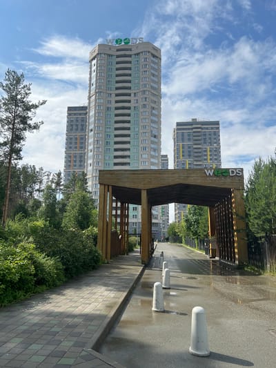 The image depicts a view of the "Woods" residential complex, located in the village of Sovkhozny, Russia. In the foreground, a wooden archway entrance is visible, with a paved walkway on the left and a wet asphalt road leading through the arch. The road is lined with white bollards. Behind the archway, several tall, multi-story residential buildings with balconies are prominent. The buildings are characterized by their light-colored facades accented with strips of mint green and yellow. The sky above is blue with scattered white clouds, and the scene is surrounded by lush green trees and foliage.