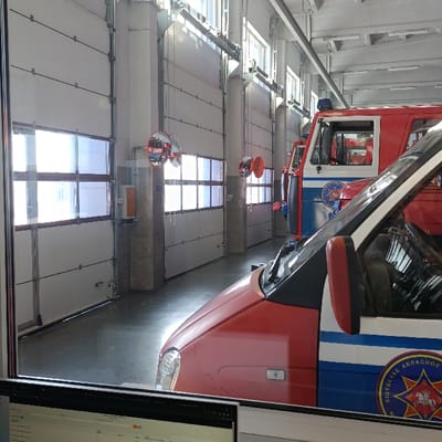The image displays the interior of an emergency vehicle bay in Hlybokaye, Belarus, viewed through a glass partition with a computer monitor partially visible in the immediate foreground.
Two emergency vehicles are parked inside the bay. In the midground, a red and blue van, identified as a GAZelle-type, features the text "ВIЦЕБСКАЕ АБЛАСНОЕ" (Viciebsk Oblast) and a star emblem with a coat of arms on its side. Behind it, a larger red and white/blue emergency vehicle, likely a fire truck, is present with a blue light bar mounted on its roof.
The bay's left wall consists of multiple white, sectional overhead garage doors, each equipped with horizontal window panels. Structural columns separate these doors, bearing mounted convex safety mirrors. Red safety cones are suspended from chains in front of some doors. The floor is a reflective, light-colored polished concrete. Overhead, exposed ceiling structures, fluorescent lighting fixtures, and high windows providing natural light are visible. No individuals or active events are depicted within the scene.