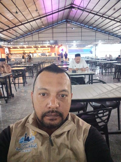 The image is a selfie taken by a man wearing a tan jacket with a logo featuring a mosque and crescent moon, and text that appears to be in Indonesian, possibly related to the city of Bireuen, Indonesia. The man is in the foreground, with his face filling a significant portion of the frame. In the background, there is a spacious indoor dining area with multiple tables and chairs. Some tables are occupied by other individuals. On the left side, a man in a patterned shirt is seated at a table. Further in the background, a man wearing a white shirt and a black cap is seated at a table looking at his phone. Behind him, a counter or bar area is visible with people working behind it. The ceiling is high with exposed beams and industrial-style lighting fixtures. The overall setting suggests a restaurant or food court.