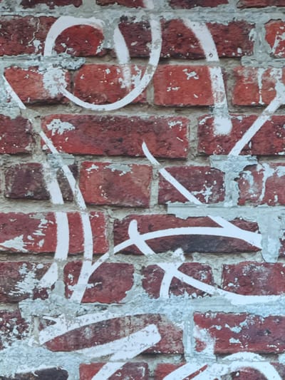 The scene depicts a weathered brick wall in Oberhausbergen, France, entirely covered with white spray paint graffiti. The wall is constructed from rectangular red bricks, showing variations in hue from dark reddish-brown to lighter orange-red. The mortar lines between the bricks are light gray and exhibit signs of cracking and flaking. The white spray paint is applied in abstract, non-figurative lines and curvilinear shapes, appearing as tags or stylized lettering that is not clearly legible. The paint application varies in density, with some areas showing solid white coverage while others are more diffuse or thin. No individuals, specific objects beyond the wall and paint, or interactions are present in the image. The environment is limited to this section of the brick wall.